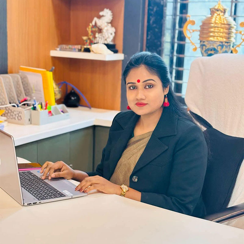 Professional woman in a blazer typing on a laptop at a tidy office desk, looking at the camera.